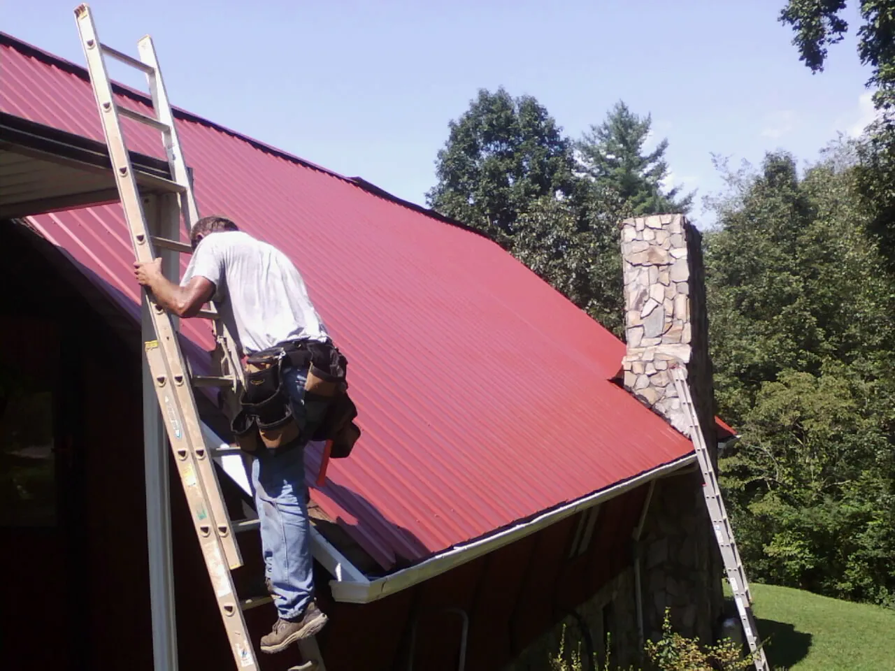 Roofer on a ladder beside a red metal roof and stone chimney — Soffit & Fascia Repair by Roofing Brothers in North Haverhill